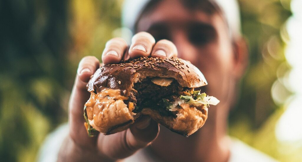 Foto van een man met een petje op zijn hoofd die een hamburger voor zich houdt waar een hap uitgenomen is.