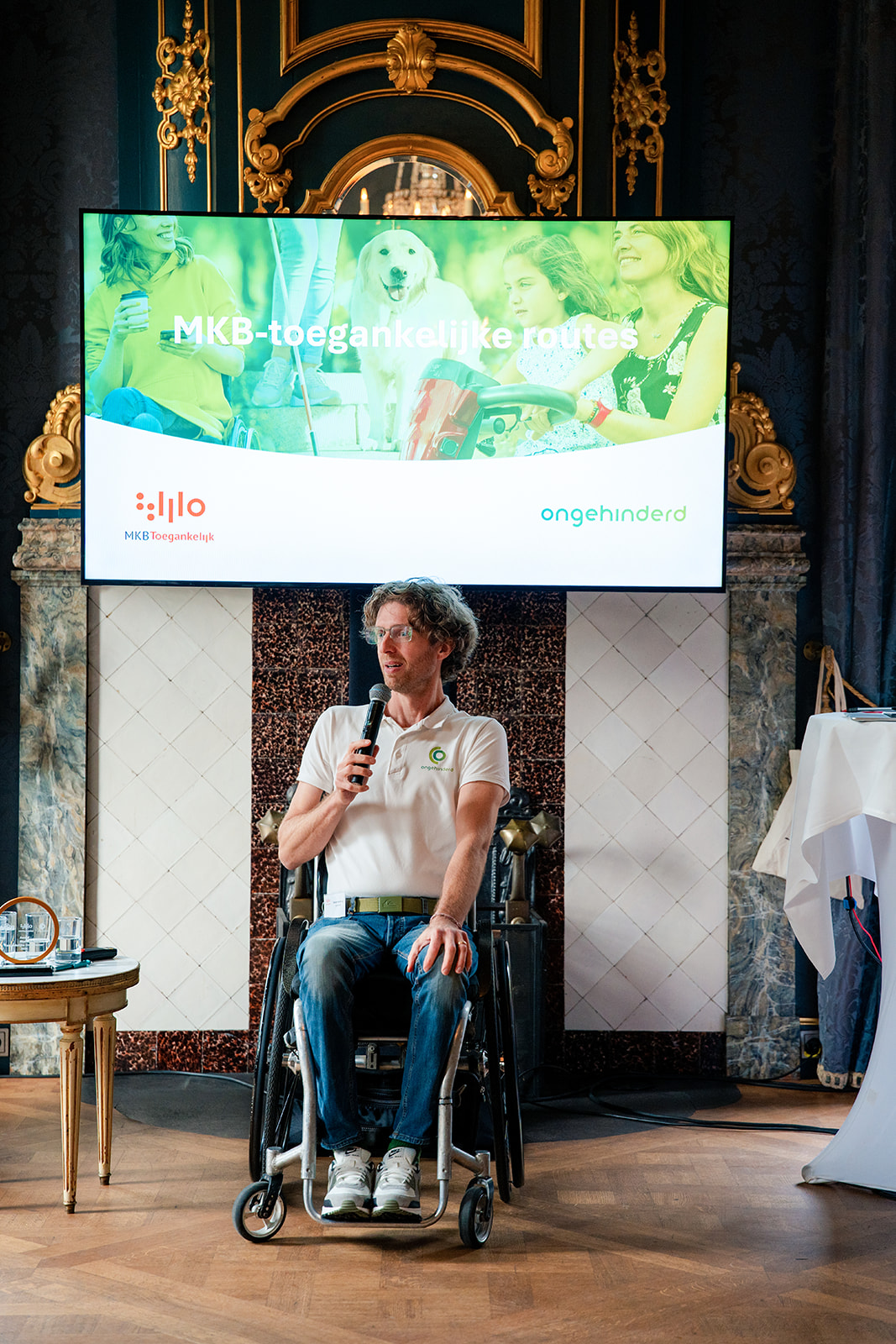 Gerard de Nooij zittend n een rolstoel. Hij heeft een microfoon in zijn handen. Achter hem is een groen gekleurde presentatie op een beeldscherm aanwezig.