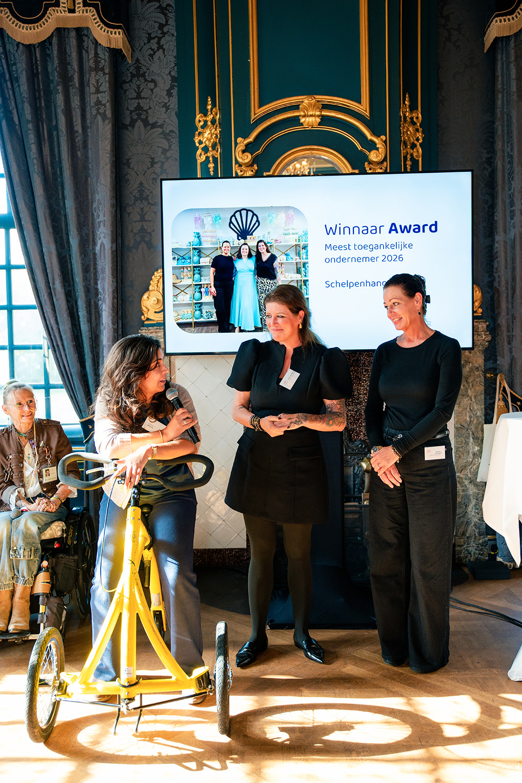 Uitreiking van de award. Links staat een dame met donker haar en gele loopfiets. Rechts staan twee dames in het zwart gekleed die de award in ontvangst nemen.